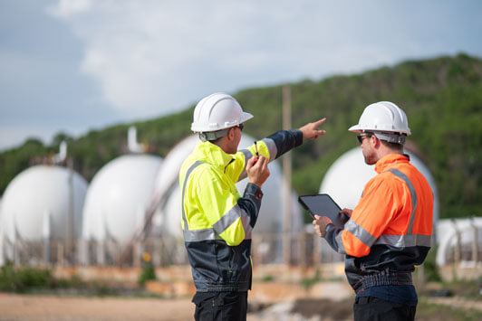 Engineers in safety gear reviewing plans near large industrial storage tanks at an energy facility