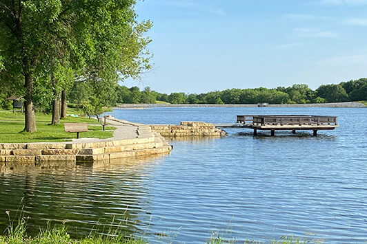 Lakeside park with walking path, trees, and a wooden observation deck over calm water