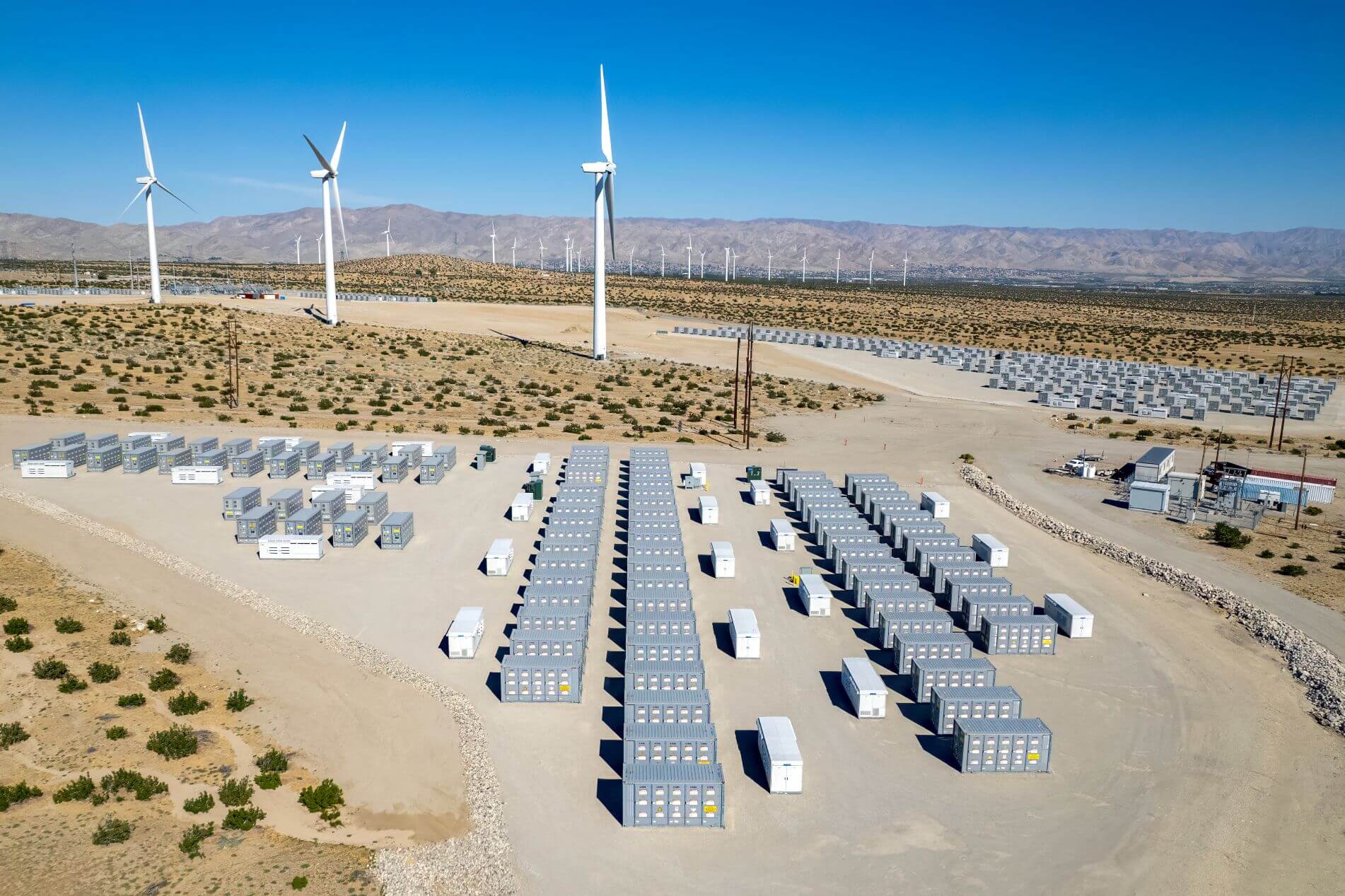 Rows of industrial battery storage units arranged in a desert near multiple wind turbines, with mountains in the background under a clear blue sky